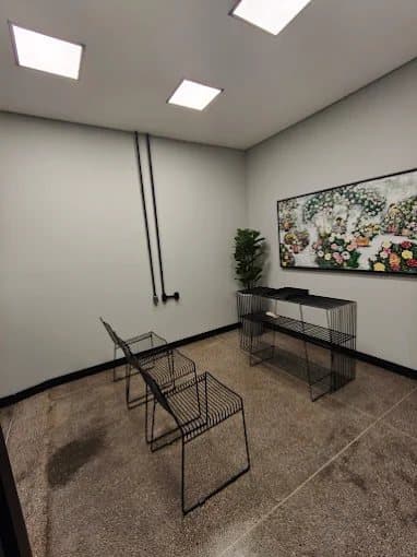 Sala de casamentos, com mesa de estrutura metálica, duas cadeiras, um quadro na parede e luminárias embutidas no teto.